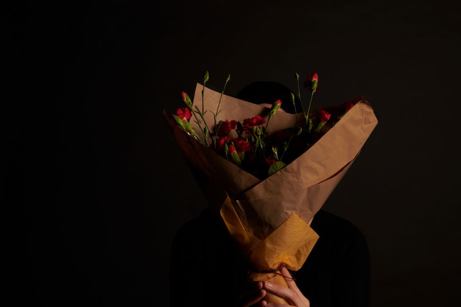 A person holding a bouquet of fresh red and pink carnations wrapped in brown kraft paper and a light brown tissue paper, with the flowers partially covering their face and head. The bouquet is positio