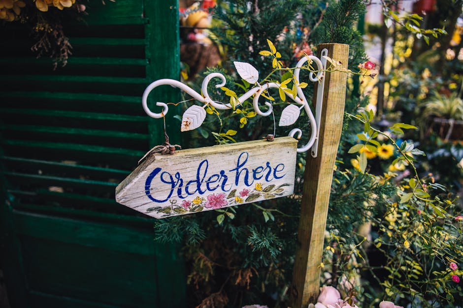 A rustic wooden sign with the word 'Order here' painted in blue cursive lettering, hanging from a decorative white metal bracket adorned with intricate scrollwork. The sign is attached to a weathered 