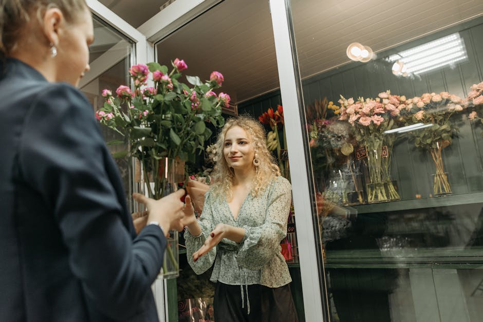 A woman with curly blonde hair and a floral blouse is receiving a bouquet of pink roses from a florist through the glass window of a flower shop. The florist, dressed in a dark jacket, is handing over