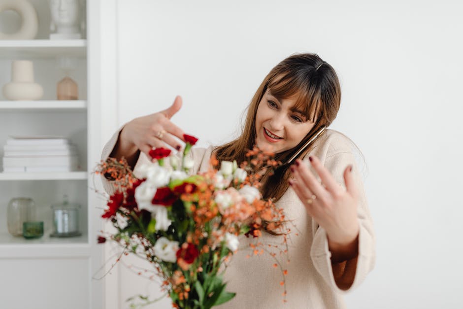 A woman with shoulder-length brown hair and a friendly expression is holding a colorful bouquet of fresh flowers, which include red roses, white lilies, pink and orange blossoms, and green foliage. Sh