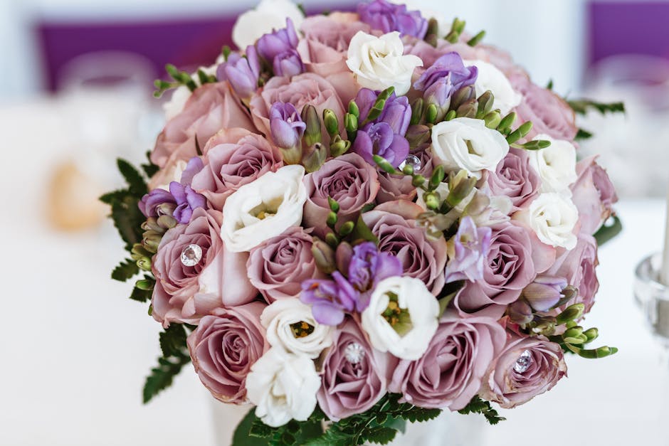 A close-up of a floral bouquet featuring soft pastel pink roses, white lisianthus, and light purple freesias, arranged in a rounded style with visible green foliage accents. The roses and lisianthus h