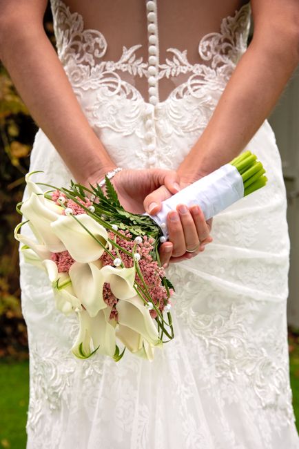 A bride wearing an elegant white gown with intricate lace and pearl buttons, holding a floral bouquet composed of white calla lilies, pink astilbe, and greenery, wrapped in white satin ribbon. The bou